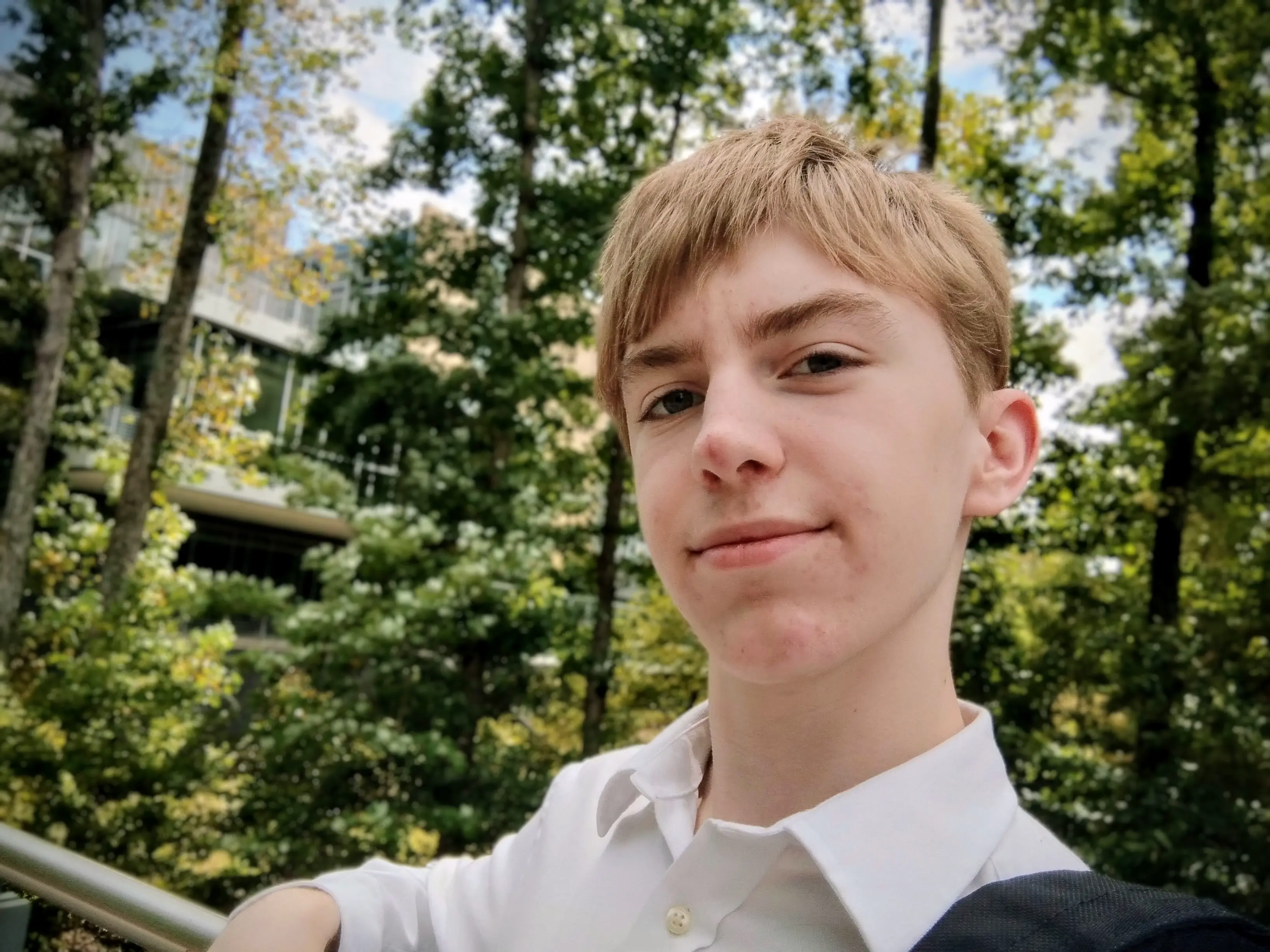 Ethan, a fair-skinned brown-haired teenager, wearing a white-collared button-up shirt, gently smiling at the camera while leaning against a railing. A vibrant, slightly blurred forest is visible in the background