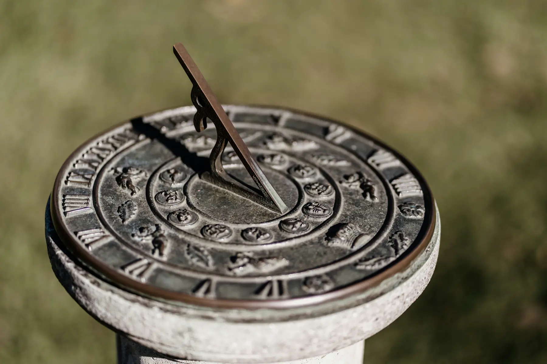 A metal sundial on a stone pedestal. On the outer circumference lie Roman numerals counting up to 24. On the inner circumference are ornate symbols. In the center is a sleek angled gnomon. The gnomon's shadow lies approximately halfway between the I and II.